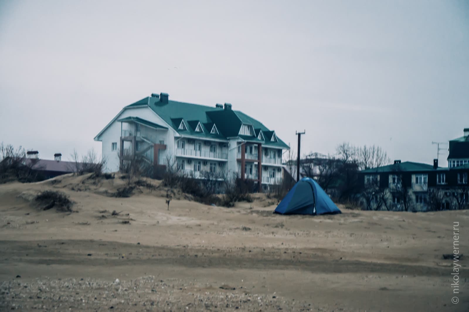 Синяя палатка среди песчанных дюн на пляже. Пасмурно. На заднем плане стоит трехэтажная гостиница.